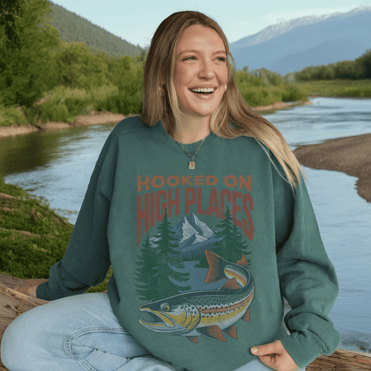 Woman wearing Hooked on High Places trout crewneck sweatshirt by a river with mountain backdrop.