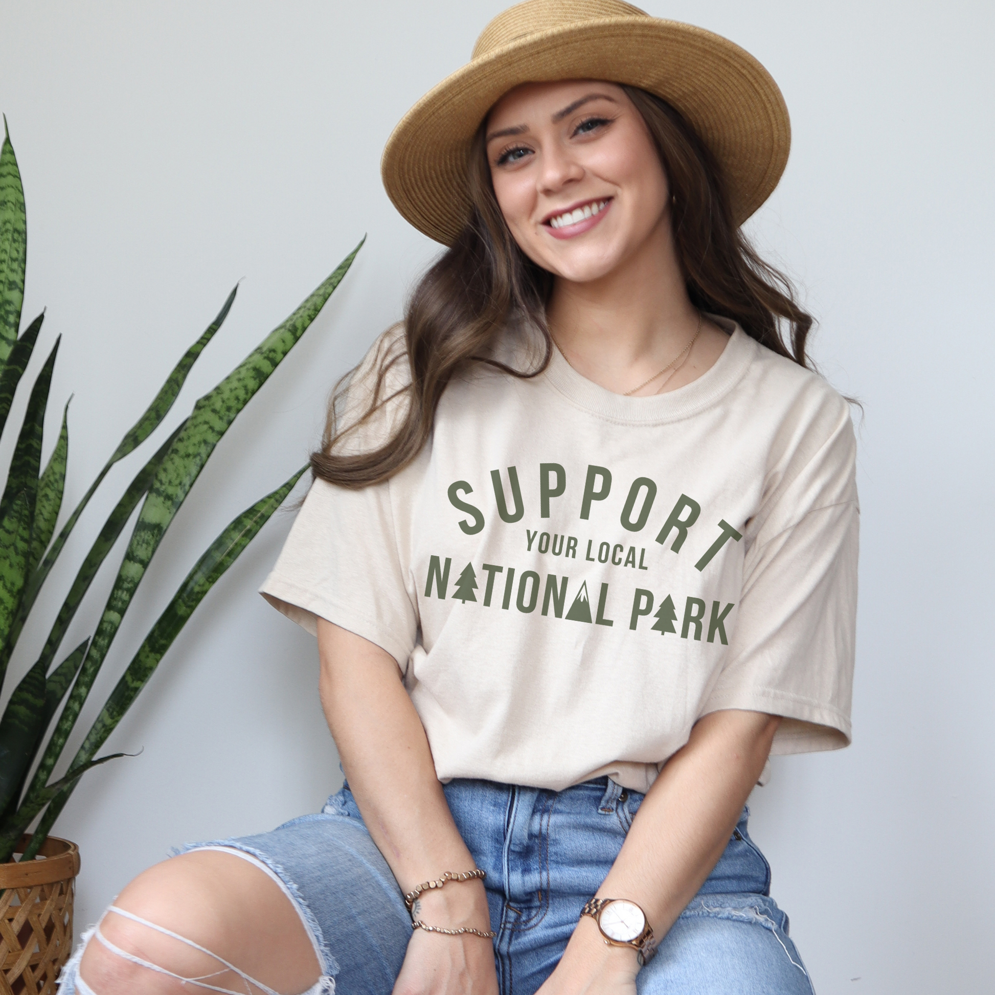 Woman wearing "Support Your Local National Park" t-shirt, smiling with a hat and greenery in the background.