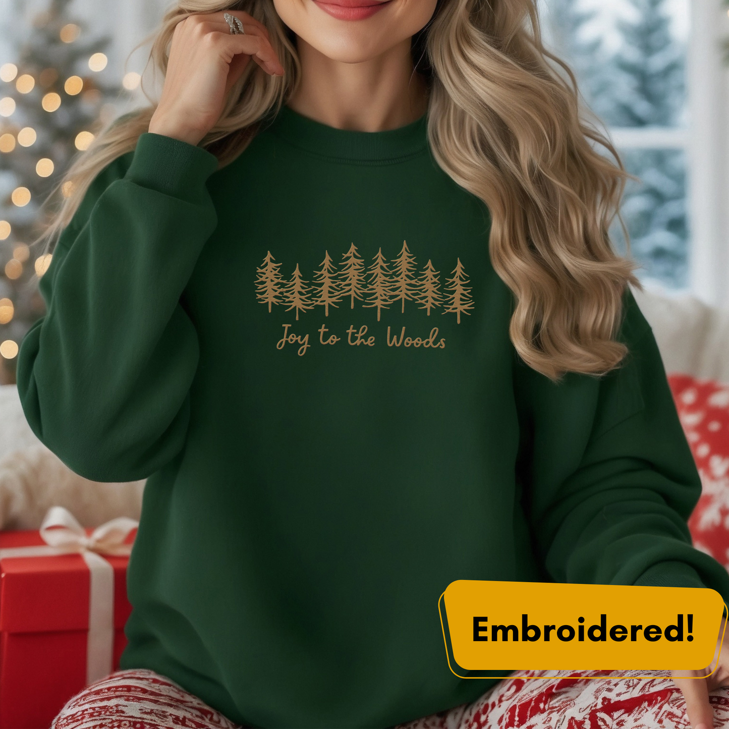Joy to the Woods embroidered crewneck in forest green with gold pine tree design and festive holiday backdrop.