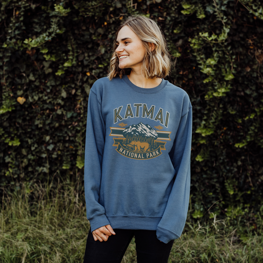 Woman wearing a cozy blue Katmai National Park sweatshirt with bear and mountain design, surrounded by greenery.