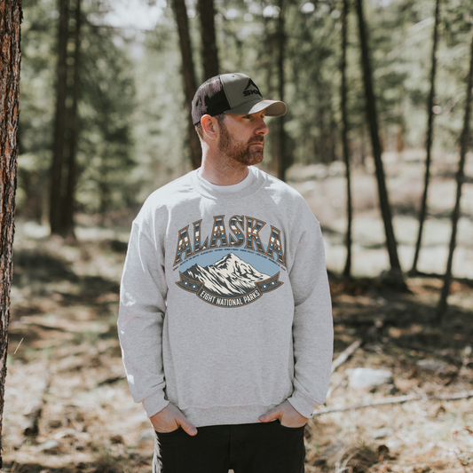 Man wearing Alaska National Parks sweatshirt in a forest setting, showcasing retro design and outdoor spirit.