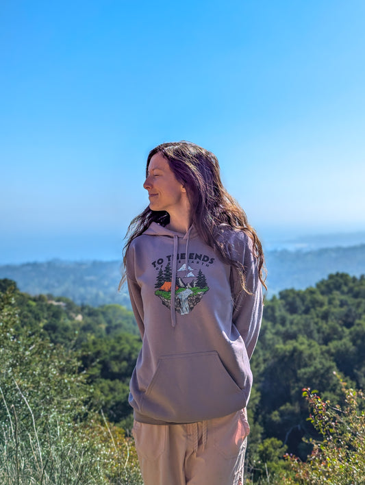Woman wearing Ends of the Earth hoodie, standing in nature with scenic hills and blue sky in the background.