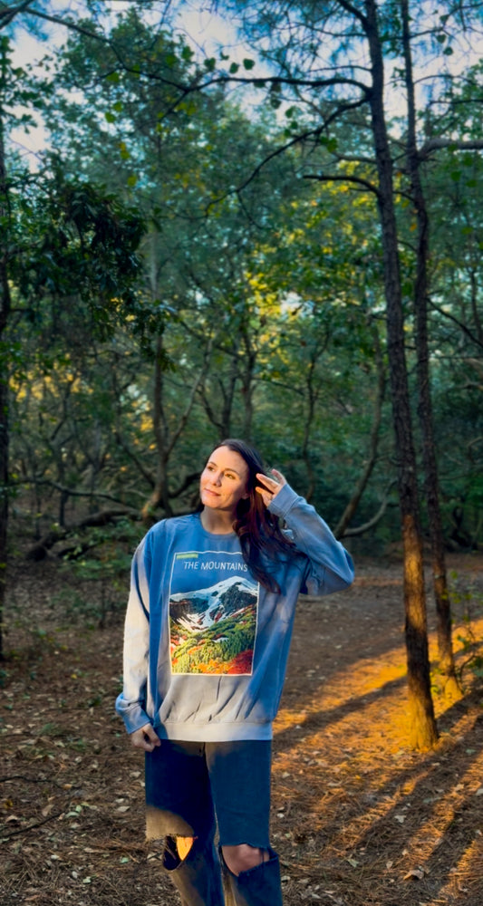 Woman standing in a forest wearing a blue sweatshirt with a colorful graphic design.