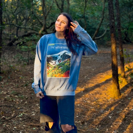Woman standing in a forest wearing a blue sweatshirt with a colorful graphic design.