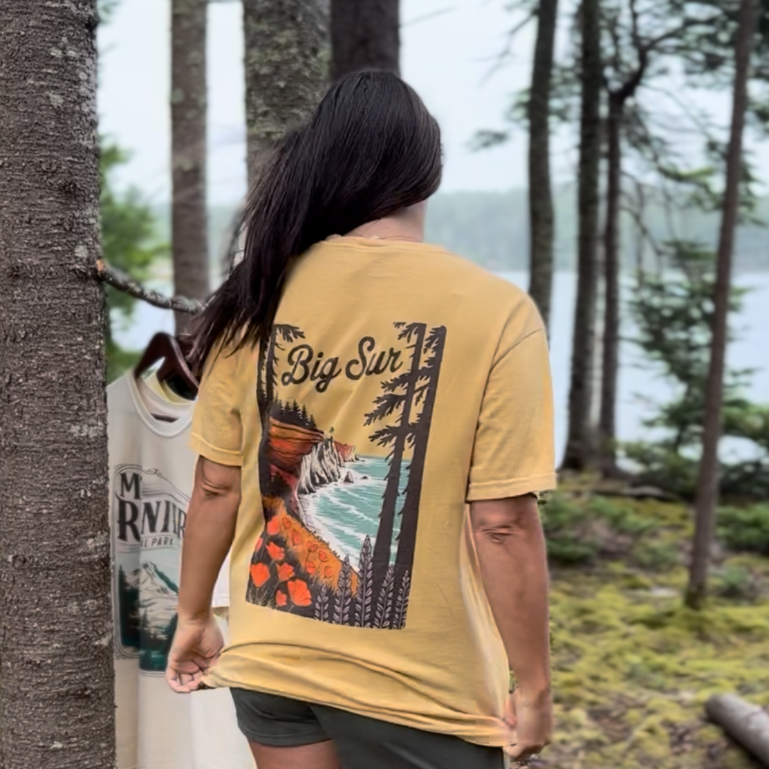 Woman wearing a vintage Big Sur shirt showcasing coastal waves and redwoods in a forest setting.