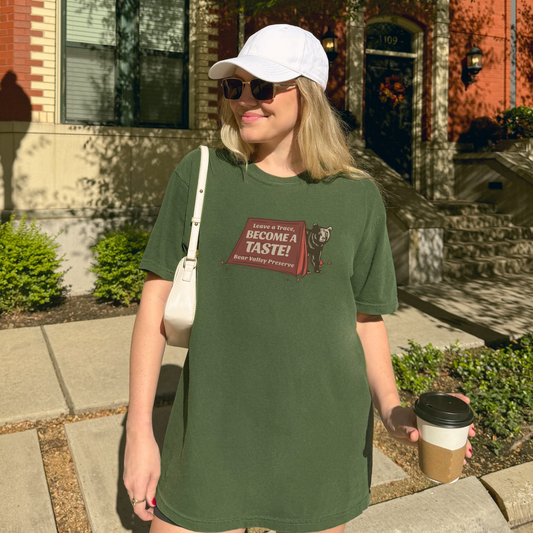 Woman wearing a green 'Leave a Trace, Become a Taste' tee with bear graphic while holding a coffee cup.
