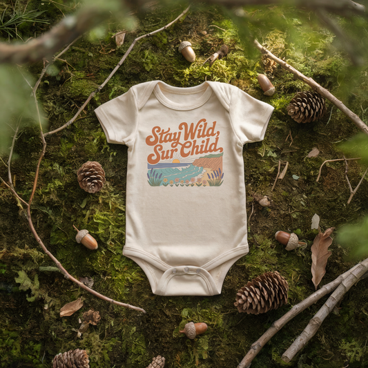 Stay Wild Sur Child baby onesie on mossy ground with pinecones, celebrating nature and adventure for little ones.