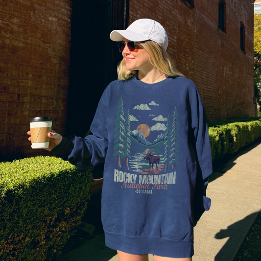 Model wearing a Rocky Mountain NP Moose Crewneck sweatshirt while holding coffee in a vibrant outdoor setting.