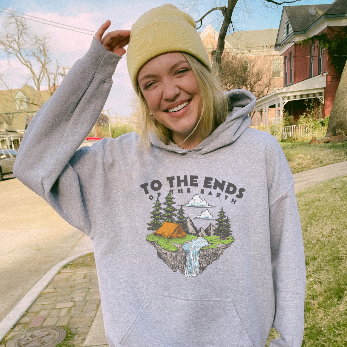Smiling woman in gray 'Ends of the Earth' hoodie with nature graphic, wearing a yellow beanie outdoors.