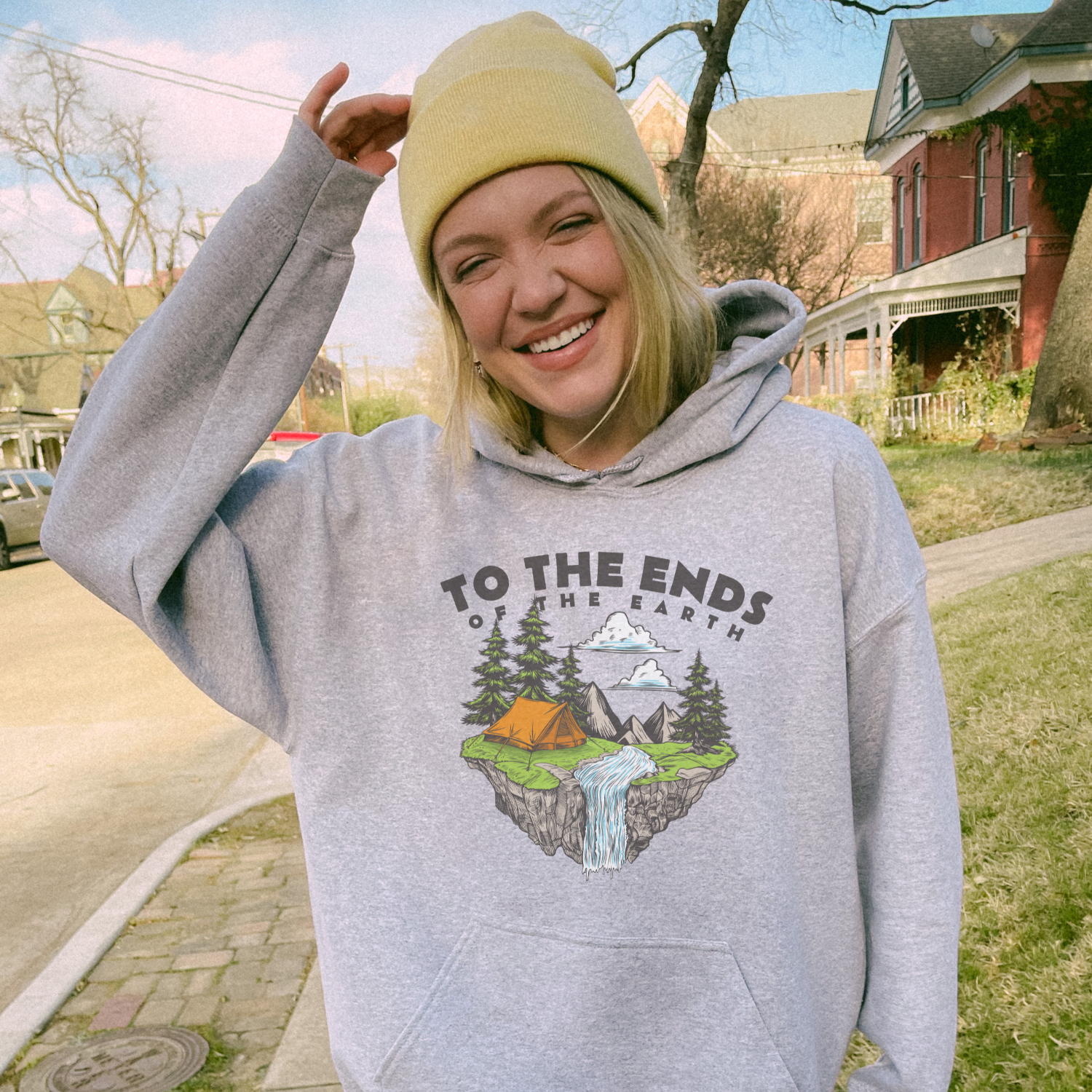 Smiling woman in gray 'Ends of the Earth' hoodie with nature graphic, wearing a yellow beanie outdoors.