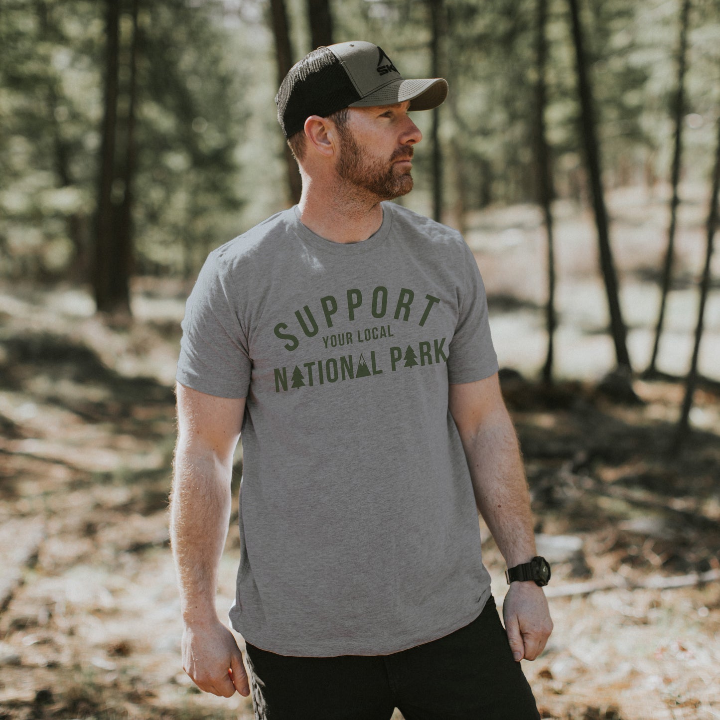 Man wearing a "Support Your Local National Park" t-shirt in a forest setting, promoting a love for nature and conservation.