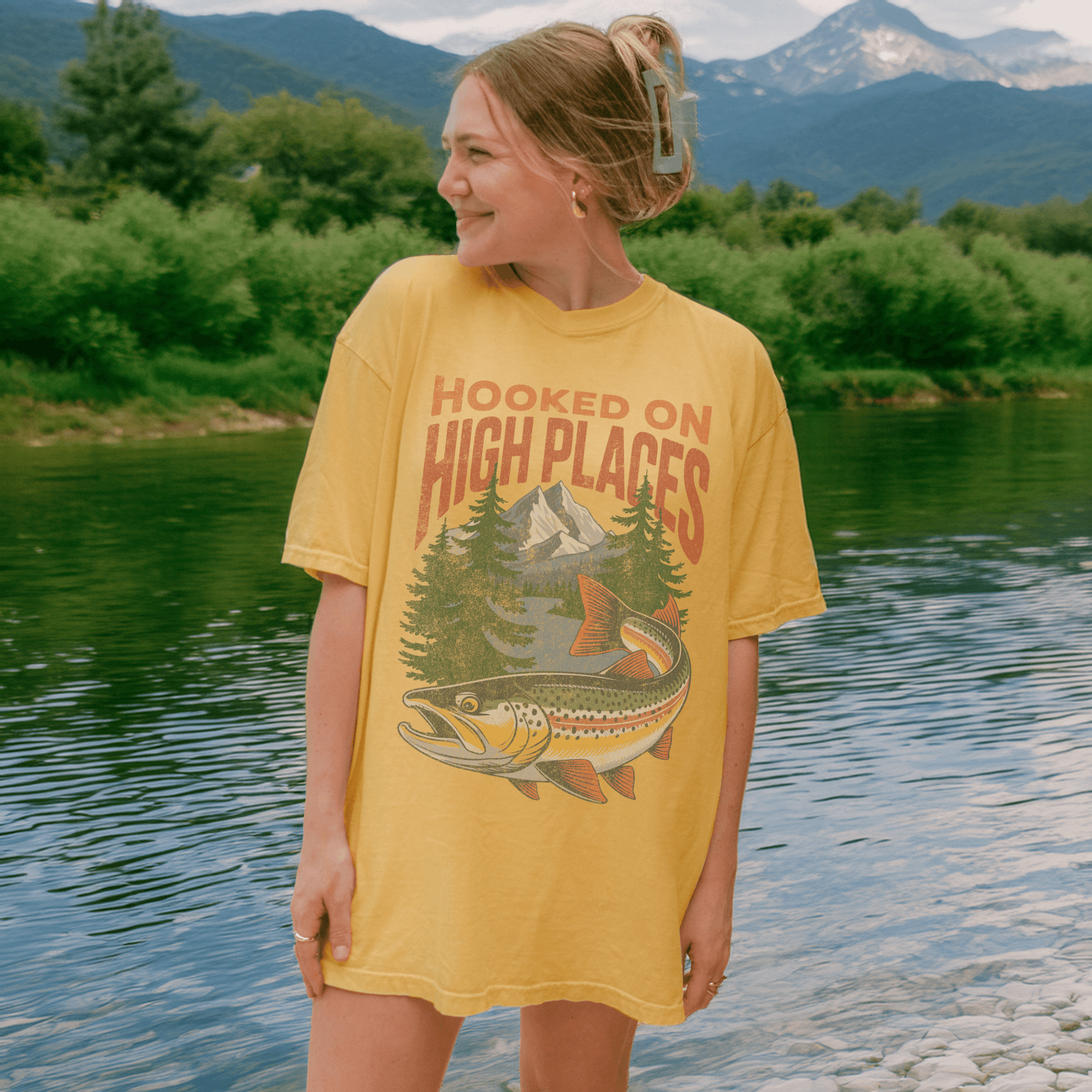 Model wearing Hooked on High Places trout adventure tee shirt by a river with mountains in the background.