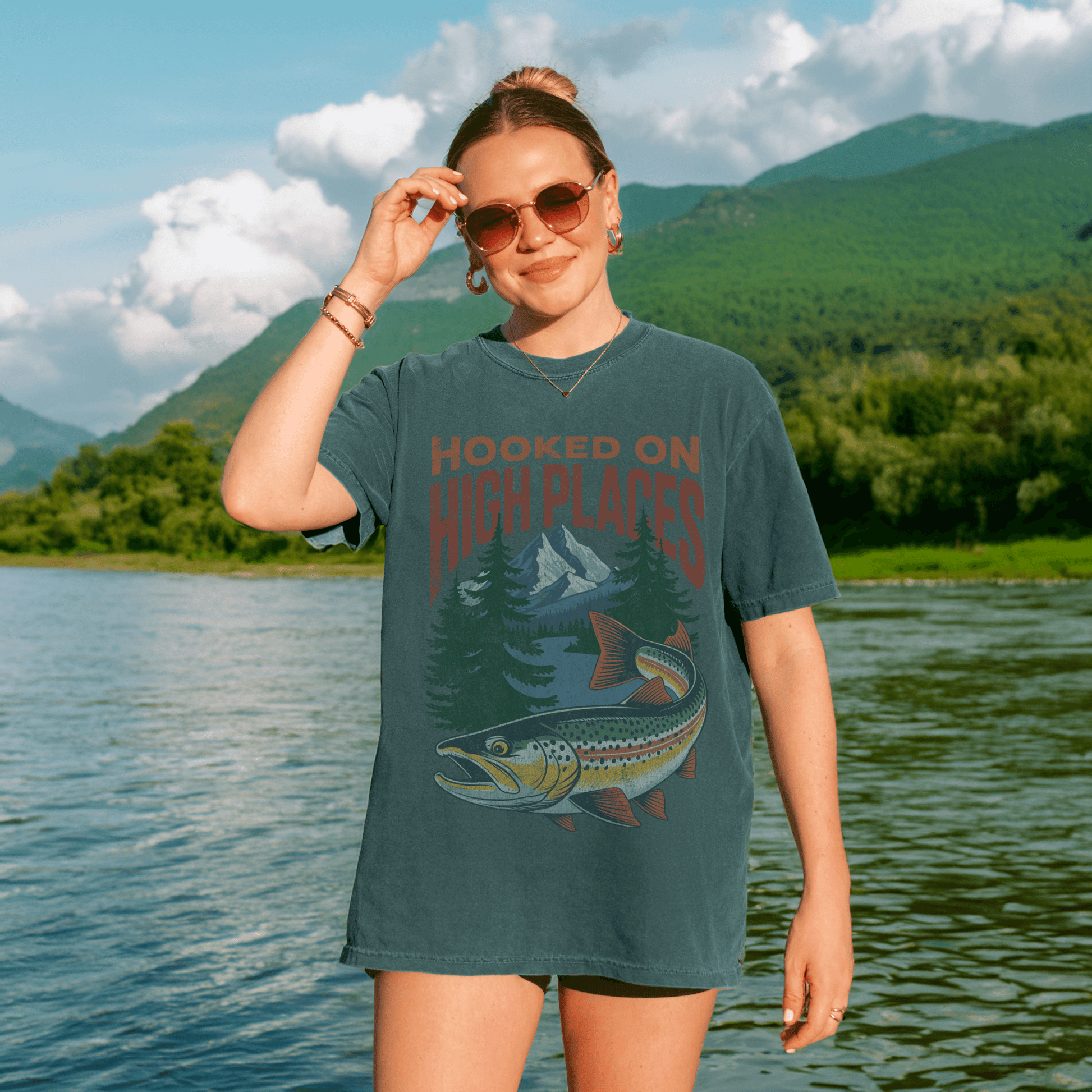 Woman wearing the Hooked on High Places trout adventure tee shirt by the river in a mountainous landscape.