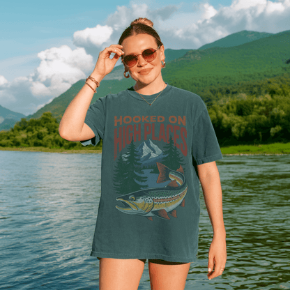 Woman wearing the Hooked on High Places trout adventure tee shirt by the river in a mountainous landscape.