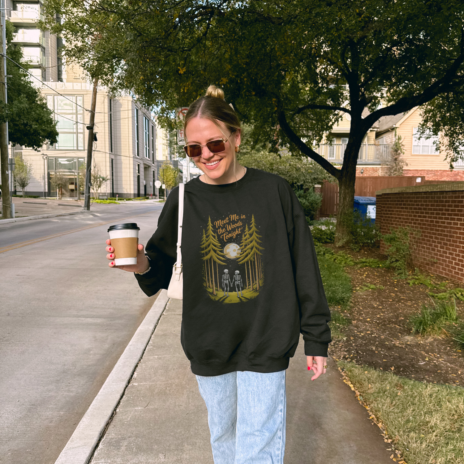 Woman wearing Meet Me in the Woods sweatshirt while walking on a city sidewalk with a coffee cup.