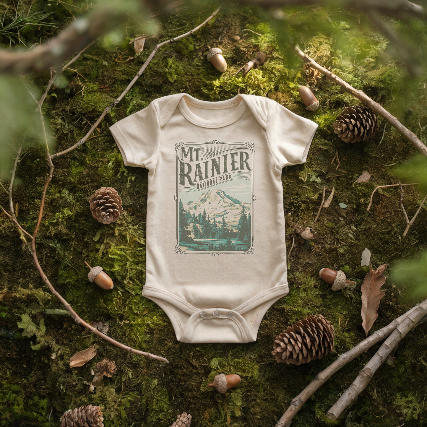 Mt. Rainier baby onesie on moss with pine cones, featuring a mountain graphic, perfect for tiny adventurers.